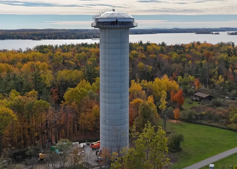 A Greatario water tower stands high on the skyline with fall coloured leaves around and a lake in the background