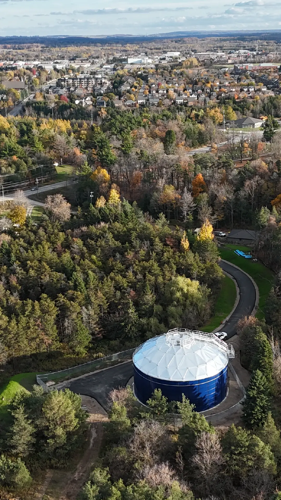 Orangeville-Greatario 1 Aerial view of a forested park with a large blue circular water tank, Orangeville Potable Water Project, in the foreground and suburban houses in the distance.