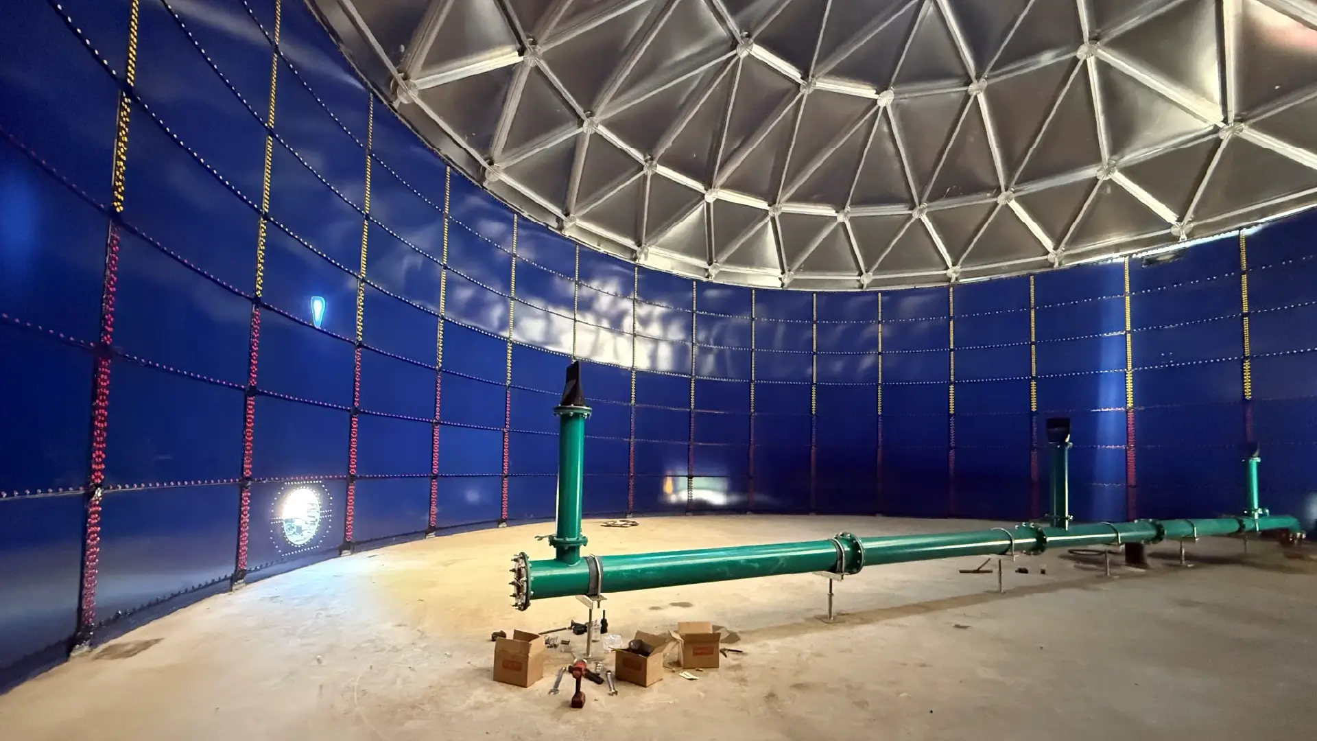 Untitled-13 Interior of a large blue-walled industrial chamber with a metal lattice dome and green pipes along the floor, tools scattered nearby.